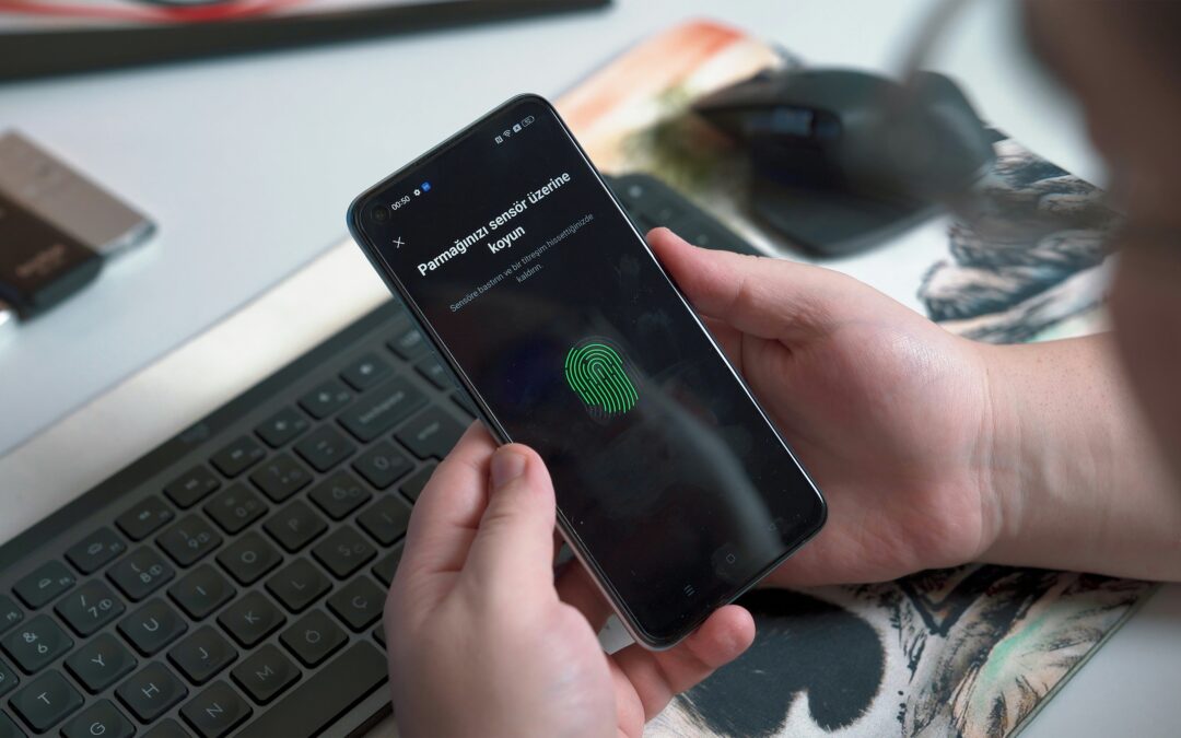 Person holding phone in two hands above a computer keyboard, on a desk. The phone screen is black with a green fingerprint image and some text in Turkish